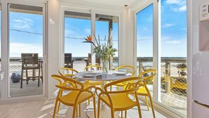 Un comedor con una mesa redonda y sillas amarillas, ventanas amplias con vista a la playa y una decoración floral en el centro.