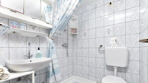 A modern bathroom with a white sink, toilet, and shower area. There are shelves with toiletries and a towel rack.