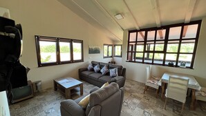 Ein großzügiges Wohnzimmer mit einer Couch, einem Couchtisch und einem Essbereich mit Tisch und Stühlen. Große Fenster sorgen für natürliches Licht und Blick auf Grünflächen.