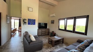 Ein geräumiges Wohnzimmer mit einer Couch, einem Couchtisch und einem Fernseher. Ein Fenster bietet Blick auf Grünflächen.