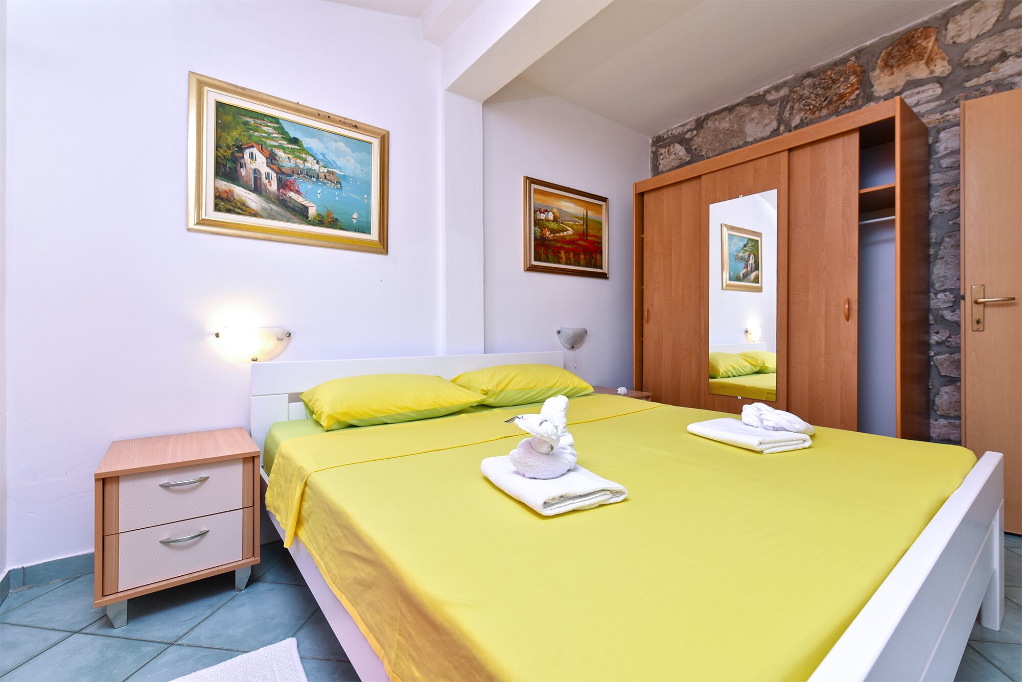 Un dormitorio con una cama, dos mesitas de noche, un armario de madera y dos cuadros en la pared.