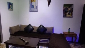 A living room with a sofa, a coffee table, and a patterned rug. There are paintings on the walls and a small table with a bottle. - CASA DAOUED APRT B (CHEFCHAOUEN)