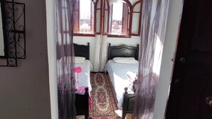 A room with two beds, a patterned rug, and a view through a window with curtains. - CASA DAOUED APRT B (CHEFCHAOUEN)