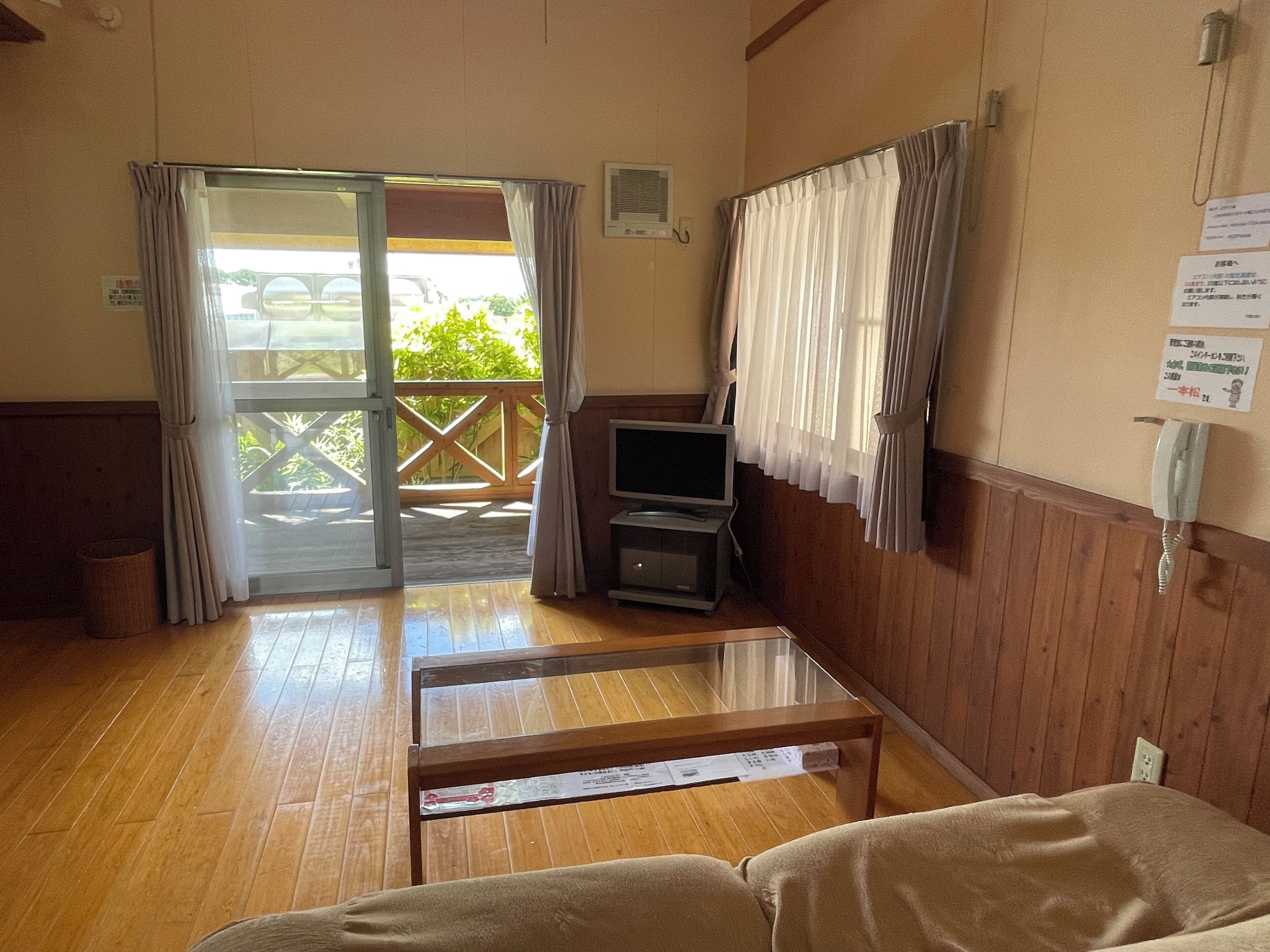 A room with a wooden floor, a sofa, a TV, a telephone, and a sliding glass door leading to an outdoor area with a wooden deck.