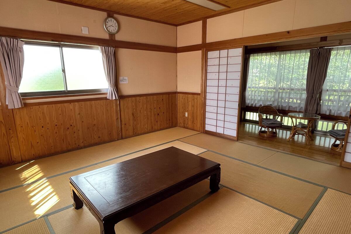 A traditional Japanese room with tatami flooring, a low wooden table, sliding doors, and a view through a window.