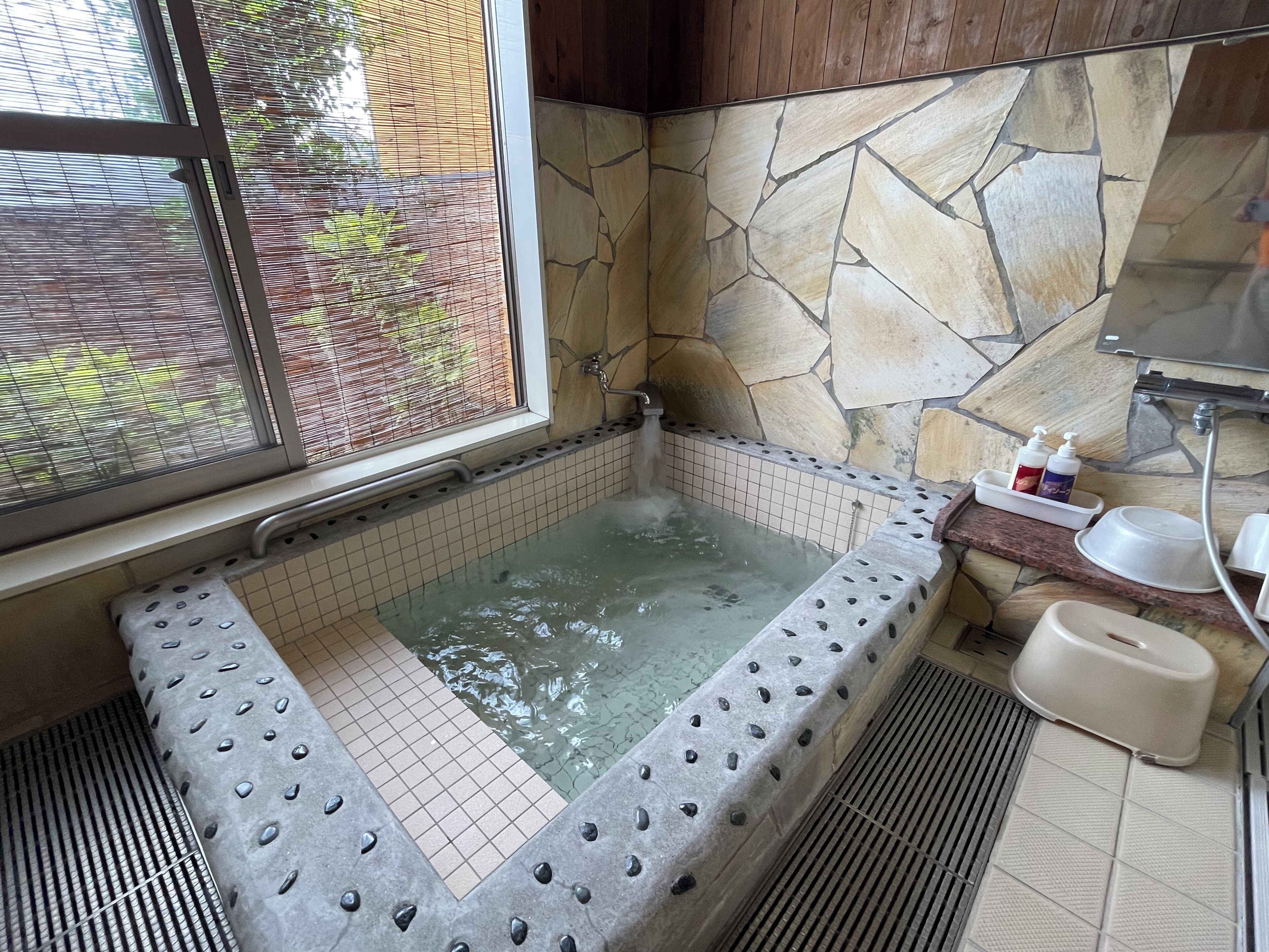 A bathroom with a stone wall, a bathtub, a window, and a shower area.
