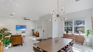 A modern living room with a large wooden dining table, comfortable seating, a ceiling fan, and a large window with a view of trees. - Tropical Oasis, Heated Pool, Near Las Olas Beach (Fort Lauderdale)