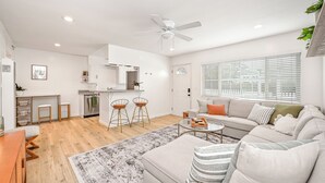 Una sala moderna con un sofá, una mesa de centro y dos taburetes altos. Se observa una zona de cocina con refrigerador, pileta y alacenas. Se distingue un ventilador de techo, y la habitación tiene un ventanal con persianas.