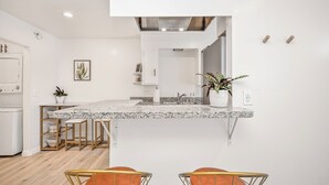 Una cocina moderna con encimera de granito, armarios blancos y campana extractora de acero inoxidable. Hay dos sillas con estructura de metal y cojines naranja.