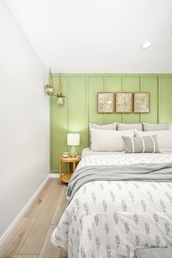 Ein Schlafzimmer mit grüner Wand, einem Bett mit weißer Bettwäsche, einem kleinen Nachttisch mit Lampe und drei gerahmten floralen Kunstwerken über dem Bett.