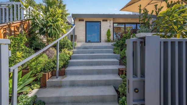 A modern house with a concrete staircase leading to a door, surrounded by lush greenery and a wooden fence.