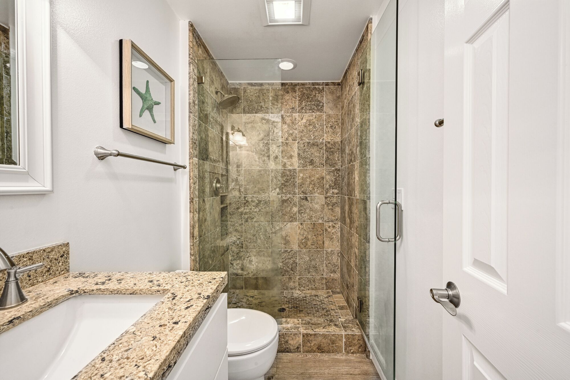 A bathroom with a granite countertop, a white toilet, a glass shower enclosure, and a door with a silver handle.