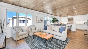 A spacious living area with a sofa set, a coffee table, and a patterned rug. There is a kitchen area with white cabinets and a dining table.
