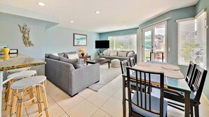 A modern living room with a grey sofa, a dining table with chairs, and a kitchen area with a bar stool. - Beachside Getaway with Patio and Parking (Newport Beach)