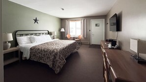 A hotel room with a bed, bedside tables, a desk, a chair, a television, and a window with curtains. - The Trail Inn - Newberry, MI - 13467 M-28 (Newberry)