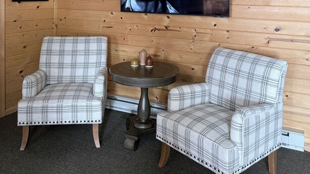 A cozy room with wooden panel walls, two plaid armchairs, a small round table, and a television mounted on the wall. - The Trail Inn - Newberry, MI - 13467 M-28 (Newberry)
