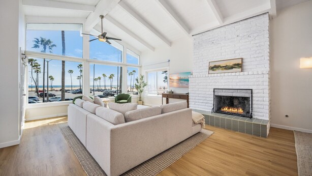 A spacious living room with a large window overlooking a beach, a white brick fireplace, and a ceiling fan. - Prime Oceanfront Location: with Direct Beach Access (Newport Beach)