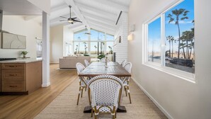 A spacious dining area with a long wooden table and wicker chairs, a kitchen with wooden cabinets, and a large window with a beach view. - Prime Oceanfront Location: with Direct Beach Access (Newport Beach)