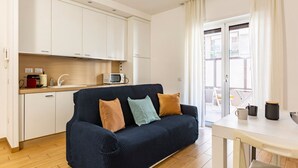 Un espacio habitable compacto con kitchenette, un sofá con cojines, una mesa de comedor y un ventanal corredero que da a un área exterior.