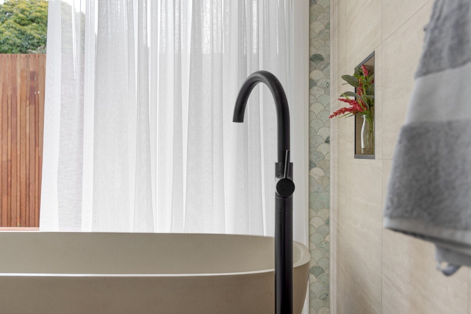Eine Badewanne mit einem schwarzen Wasserhahn, ein Holzzaun im Freien und eine geflieste Wand mit einer Blumenkomposition.