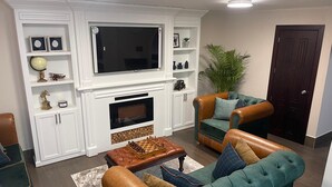 A living room with a fireplace, a television, and a chessboard on a leather ottoman.