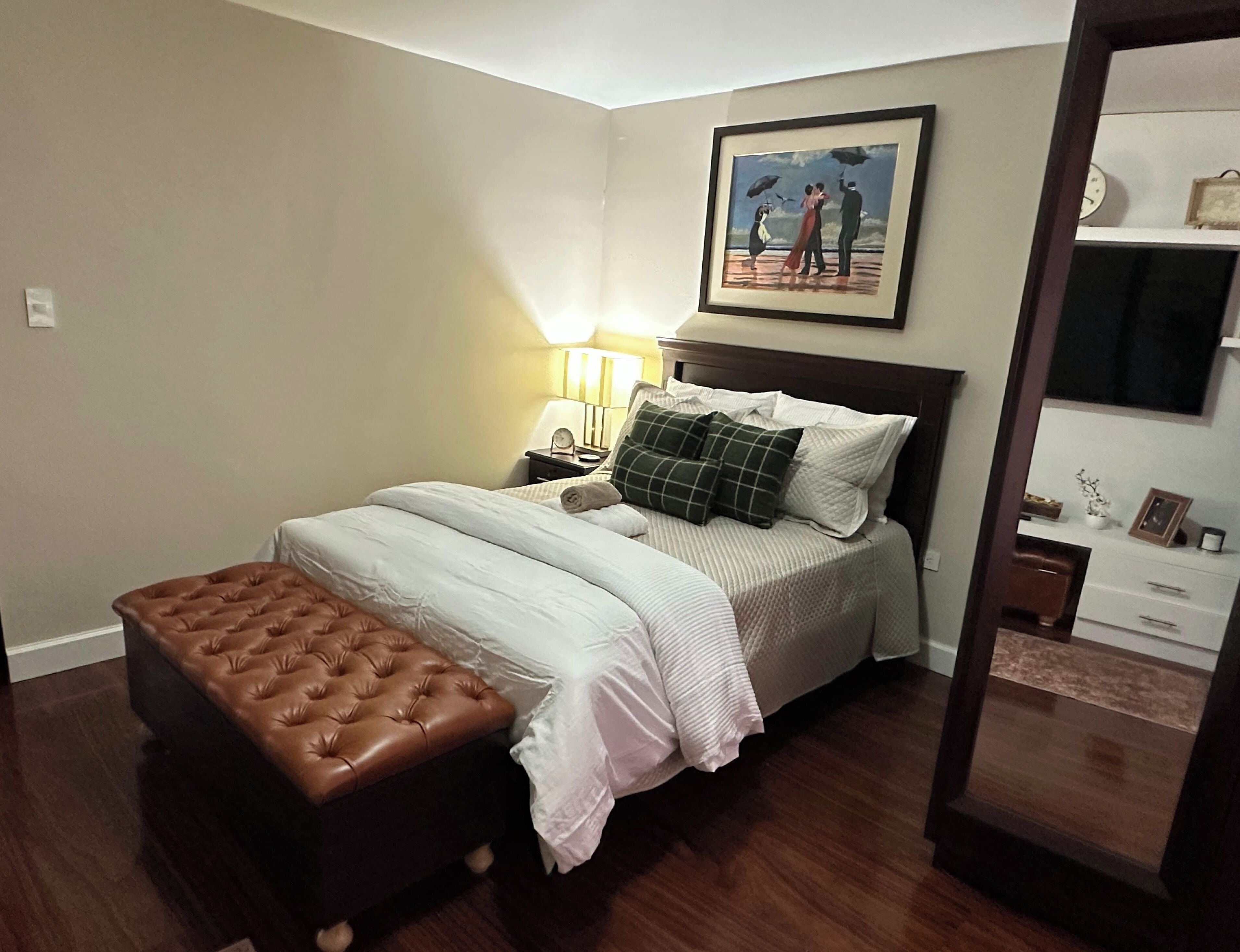 Ein ordentlich eingerichtetes Schlafzimmer mit Bett, Nachttisch, Lampe, einem gerahmten Gemälde und einem braunen Hocker.