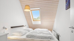 Ein Schlafzimmer mit schrägen Wänden, einem Dachfenster, einem Bett mit weißer Bettwäsche, einem Nachttisch und einer Wandleuchte.