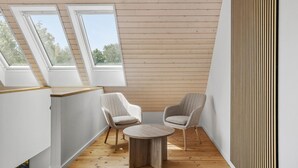 Ein modernes Schlafzimmer im Loft-Stil mit schrägter Decke, Holzboden, zwei weißen Stühlen, einem runden Holztisch und großen Fenstern mit Blick ins Freie.