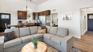 A modern living room with a sectional sofa, a wooden coffee table, and a dining area with a kitchen in the background. - New Build w/ Hot Tub, Sauna, Arcade, Mountain Views (Florissant)