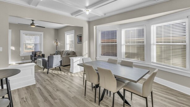 Un comedor amplio con una mesa larga y numerosas sillas, una zona de estar con sofá y sillones, y ventanas grandes con persianas.