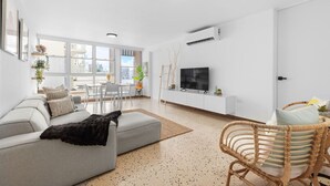 A modern living room with a grey sofa, a wicker chair, a TV, and a dining area with a table and chairs.