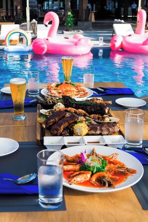A poolside dining area with a seafood platter, drinks, and pink flamingo floats. - Bounce Beach Hotel & Tien Sa Resort  (Da Nang)