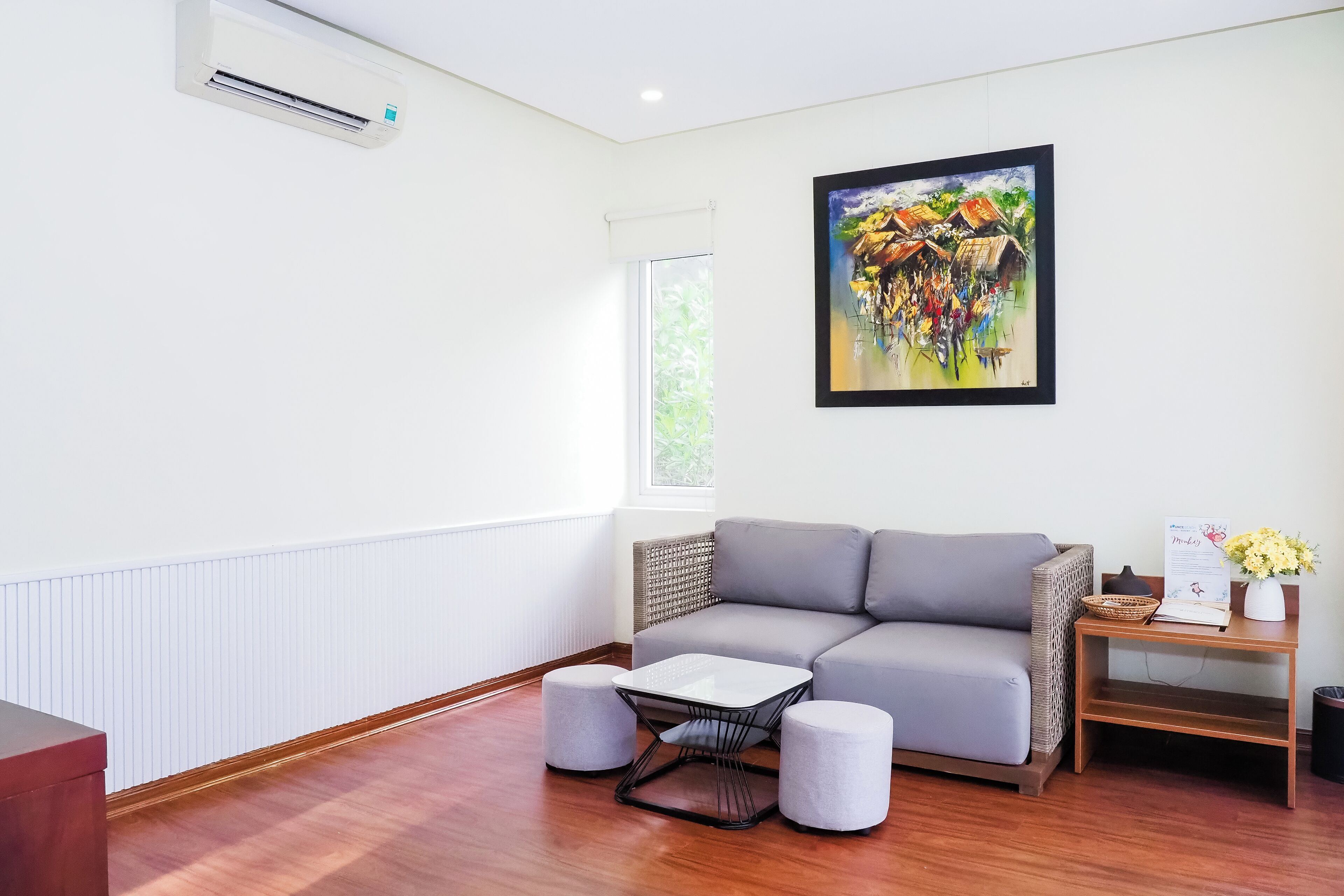 Una sala moderna con un sofá gris, una mesita blanca y dos otomanas. Un cuadro de una escena colorida cuelga en la pared, y hay una ventana que proporciona luz natural.