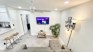 Ein modernes Wohnzimmer mit einem grauen Sofa, einem Deckenventilator, einem Flachbildfernseher, einem kleinen Holztisch und einem Topf mit Pflanze.