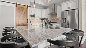 A modern kitchen with white cabinets, a stainless steel refrigerator, a marble island with bar stools, and pendant lighting.