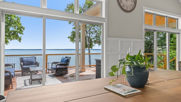 A room with a wooden table, a potted plant, and a view of a balcony with wicker furniture and a railing.