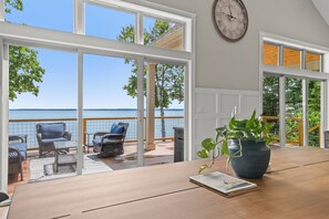 A room with a wooden table, a potted plant, and a view of a balcony with wicker furniture and a railing. - Spectacular Lakefront Retreat on Lake Michigan in Suttons Bay (Suttons Bay)