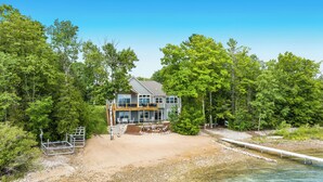 A two-story house with a deck, surrounded by dense trees and a sandy beach.