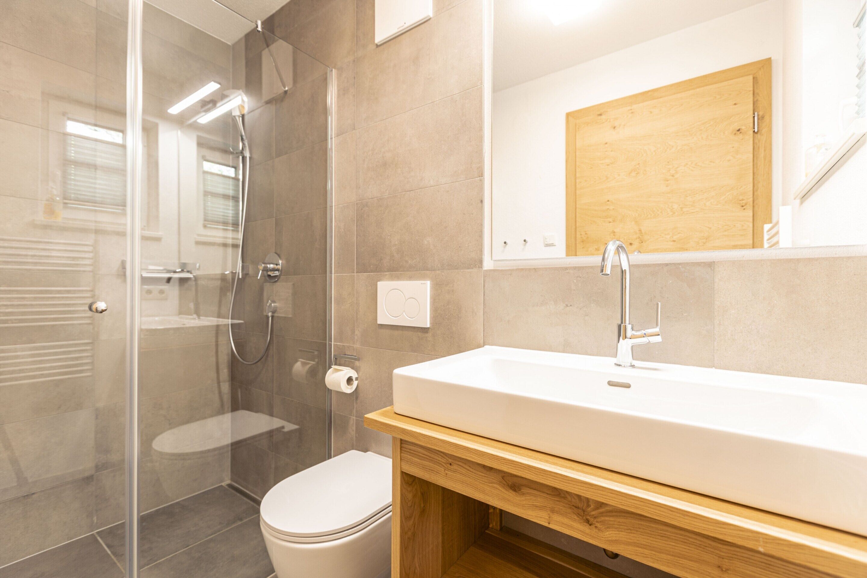 Une salle de bain moderne avec une douche à parois de verre, des toilettes blanches, un meuble-lavabo en bois et un placard en bois au-dessus du miroir.