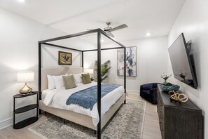 Une chambre à coucher moderne avec un lit à baldaquin, une table de chevet avec une lampe, une commode de couleur sombre et un grand miroir au mur.