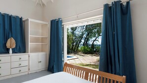 Ein Schlafzimmer mit einem Bett, einem hölzernen Kopfteil, einer weißen Kommode mit Schubladen, einem Regalsystem und Blick auf einen Garten und Bäume durch ein Fenster mit blauen Vorhängen.