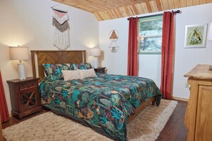 A bedroom with a bed, two bedside tables, a nightstand, a wooden headboard, a window with curtains, and wall decorations.