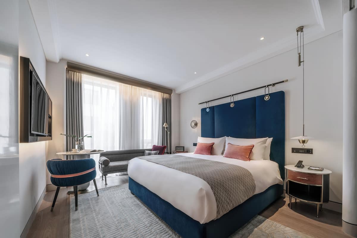A modern hotel room with a large bed, a desk, a chair, and a view of the outdoors.