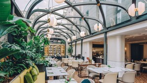 Un restaurant avec de hauts plafonds, des luminaires suspendus et des plantes d’intérieur. Les tables sont dressées avec des nappes blanches et des serviettes vertes.