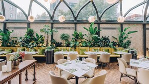Un restaurant doté d’un plafond haut et de grandes fenêtres, où les tables sont dressées avec des nappes blanches et des serviettes vertes, et où l’on trouve de nombreuses plantes vertes.