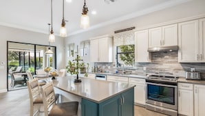 A modern kitchen with a central island, stainless steel appliances, and a view to the outdoor patio. - 12894 Epping Way (Fort Myers)