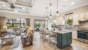 A spacious living area with a sofa, coffee table, and dining table, connected to a kitchen with a large island and modern appliances. - 12894 Epping Way (Fort Myers)