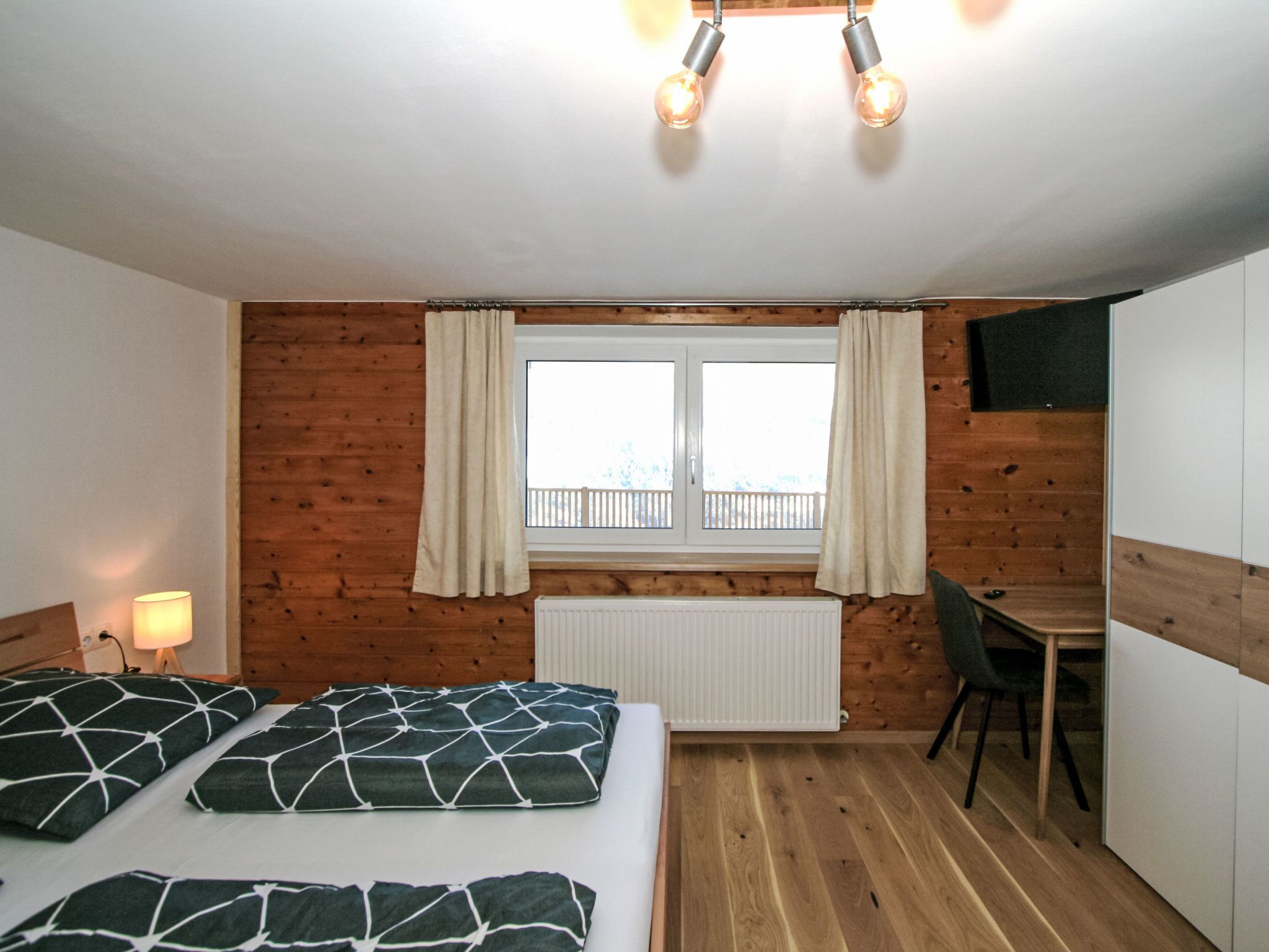 A bedroom with two beds, a window with curtains, a radiator, a desk, and a chair.
