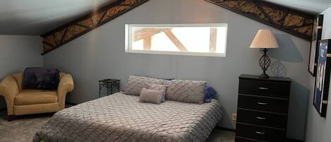 Ein Schlafzimmer mit schrägem Dach, einem Bett, einem Sessel, einer Kommode und einem Fenster.