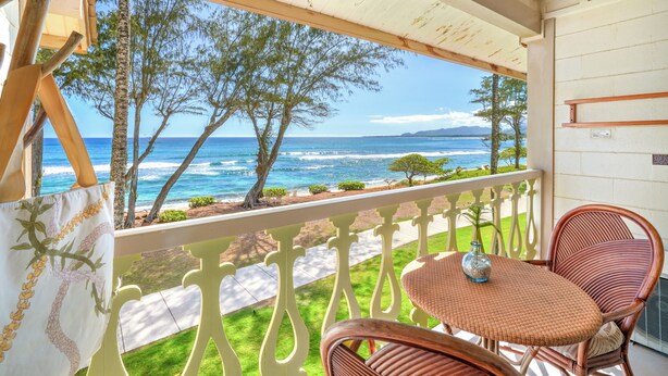 Een balkon met rieten stoelen en een tafel, met uitzicht op een strand met bomen en de oceaan.